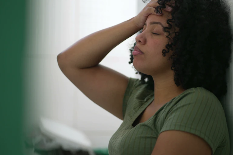 A woman with her eyes closed and an exhausted expression on her face holds her hand to her forehead and leans against a wall