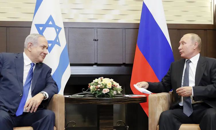 Russian president Vladimir Putin and Israeli prime minister Benjamin Netanyahu sit looking at each other with Russian and Israeli flags behind them.
