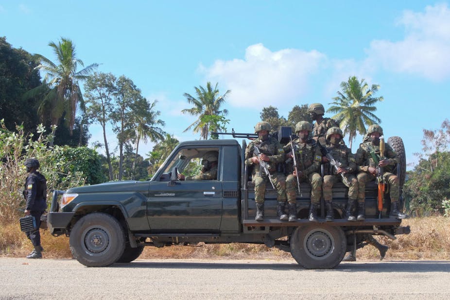 A group of soldiers in green fatigues, helmets and face masks sitting on the back of a pickup vehicle holding guns with the barrels facing the ground