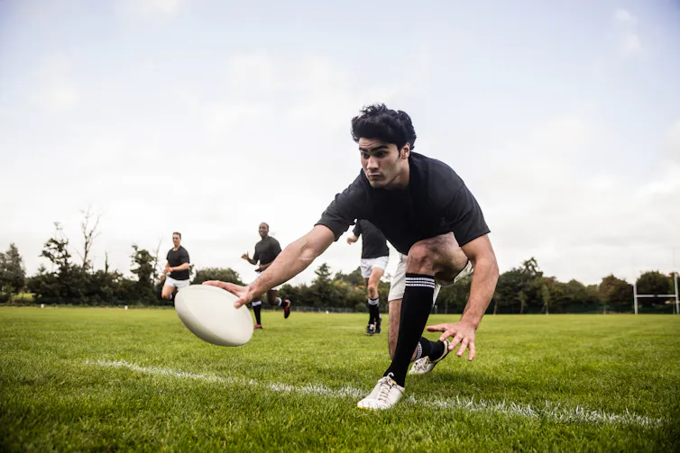 A man playing rugby bends down to place the ball on the ground to score a try.