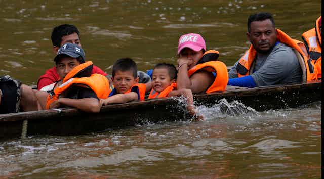 A boat is low in the water carrying two children, one smiling, and several adults. All are wearing orange life jackets.