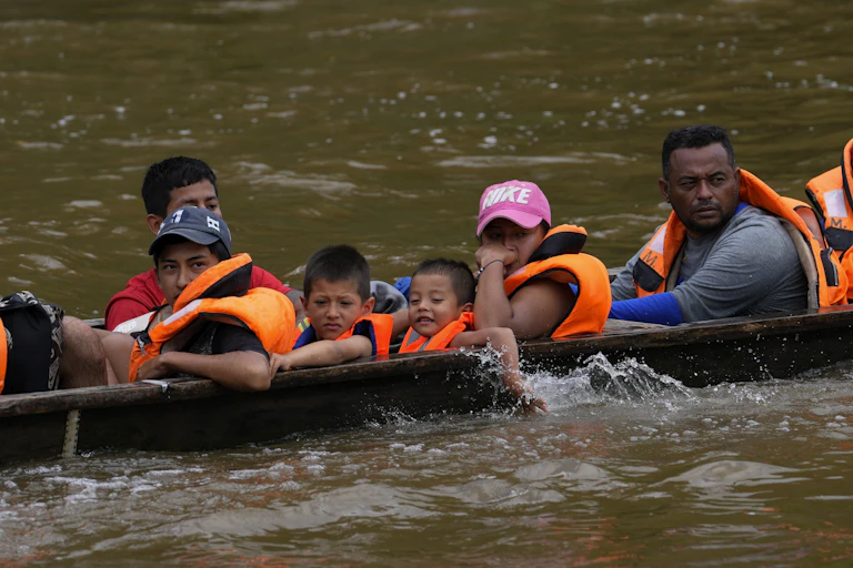 A boat is low in the water carrying two children, one smiling, and several adults. All are wearing orange life jackets.