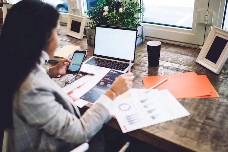 A young woman sitting at a desk with financial papers, looking at her laptop and mobile phone