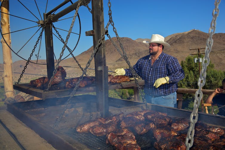 Man wearing a large cowboy hat cooks beef on outdoor grills under a clear blue sky.