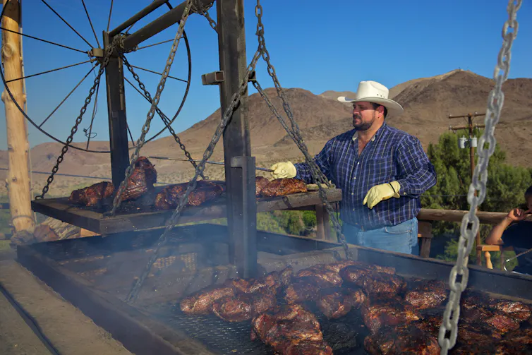 Man wearing a large cowboy hat cooks beef on outdoor grills under a clear blue sky.