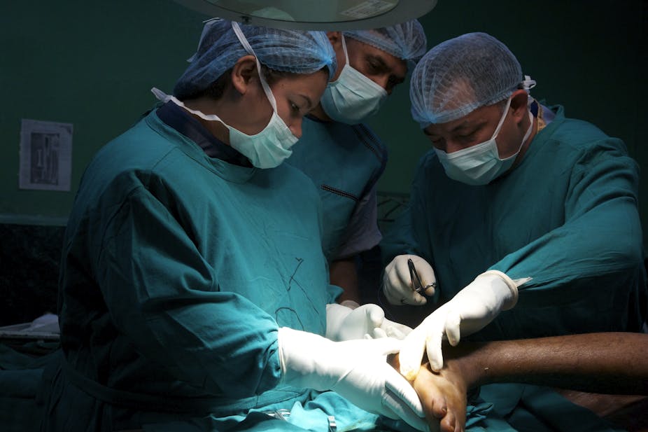 Trois soignants en blouse, portant des masques chirurgicaux et des charlottes sur la tête, sont en train d'opérer une personne blessée au pied.