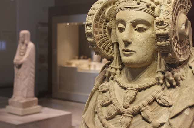 Un busto de piedra de una mujer engalanada en primer plano con otra escultura de piedra de una mujer engalanada en el fondo, ambos en la sala de un museo.