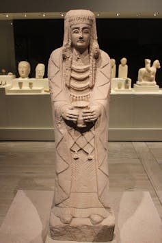Estatua en piedra de una mujer de pie, engalanada, situada en la sala de un museo.
