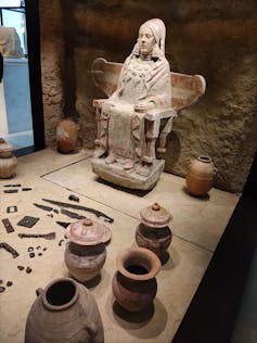 Estatua de una mujer en piedra, sentada, rodeada de vasijas y armas.