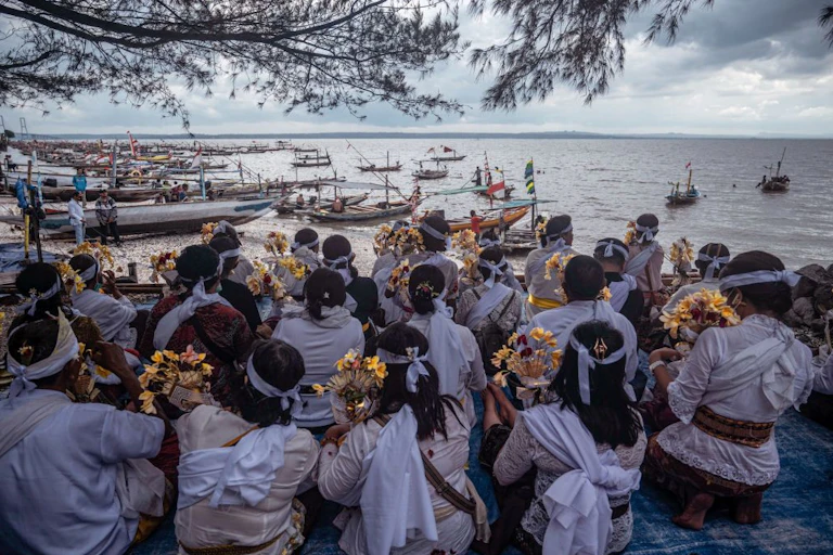 Around two dozen people wearing white face the shoreline where many small boats are tied up.