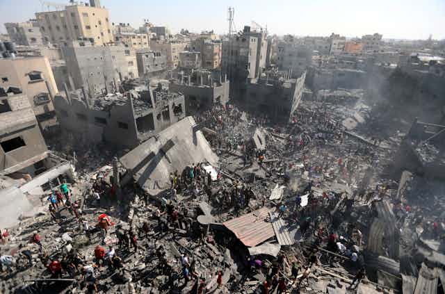 Photo of devastation following an airstrike on the Gaza Strip, with collapsed and burned buildings and crowds of people inspecting the rubble for survivors.