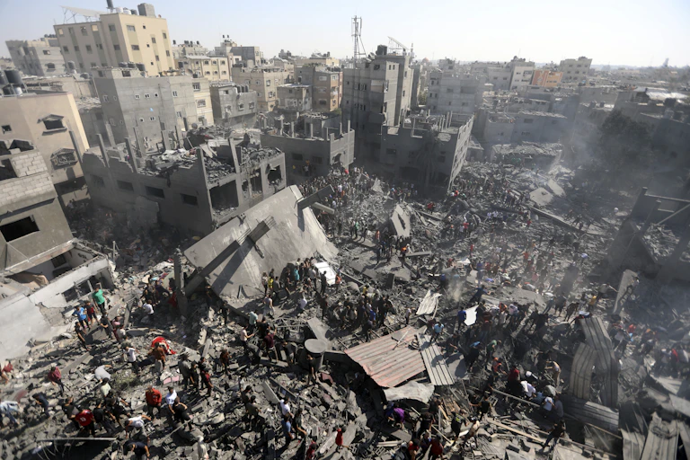 Photo of devastation following an airstrike on the Gaza Strip, with collapsed and burned buildings and crowds of people inspecting the rubble for survivors.