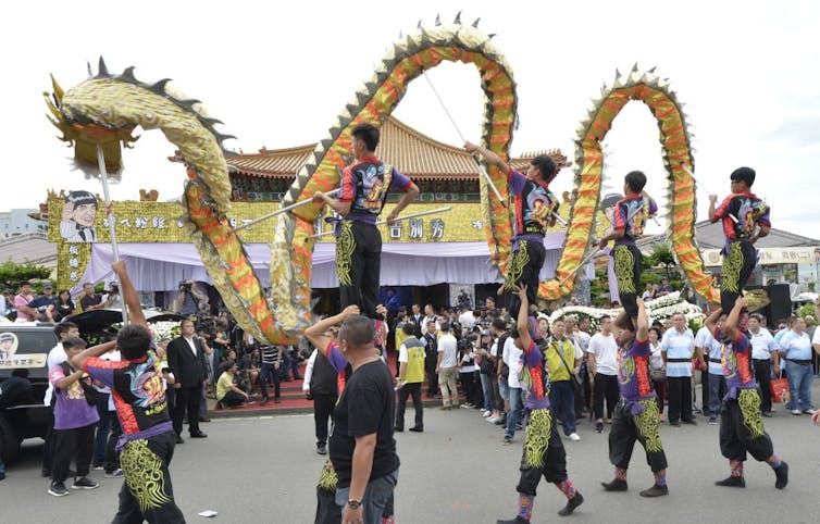 Nine men in black outfits with brightly colored patterns on them hold a huge puppet of a dragon outside a building with Chinese characters on it.