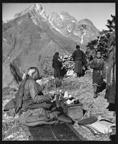 A black and white photo shows a man seated in prayer on top of a mountain, as other people work in the background.