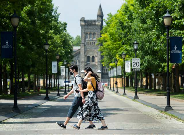 Two people wearing face masks cross a tree-lined street in front of a tall, stone building