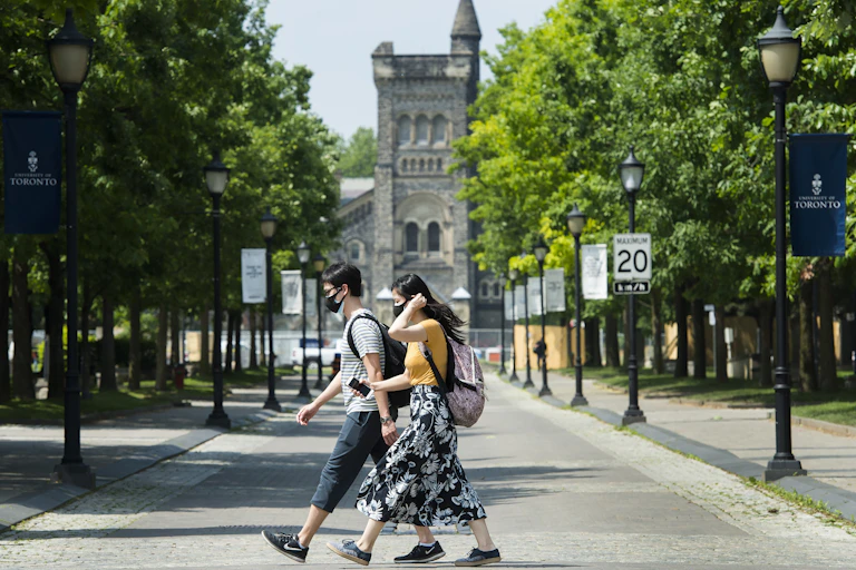 Two people wearing face masks cross a tree-lined street in front of a tall, stone building