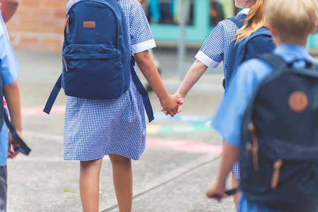 Children in their school uniforms are walking toward their school wearing backpacks and two of them are holding hands.