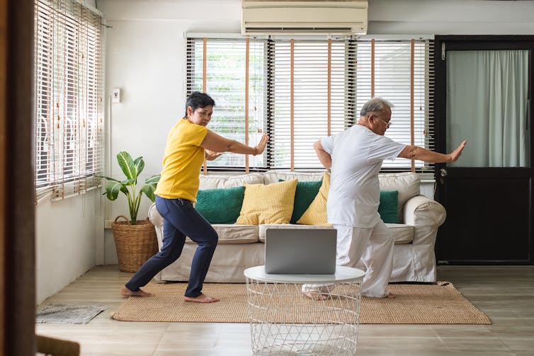 An elderly couple performs tai chi in their home.