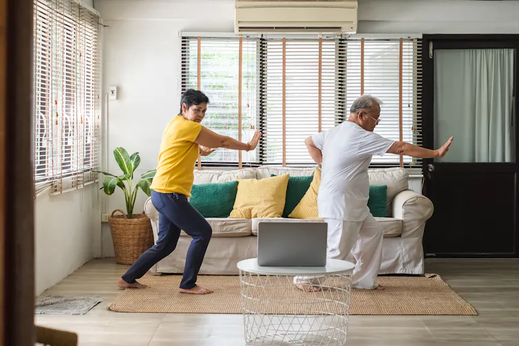 An elderly couple perform tai chi in their home.