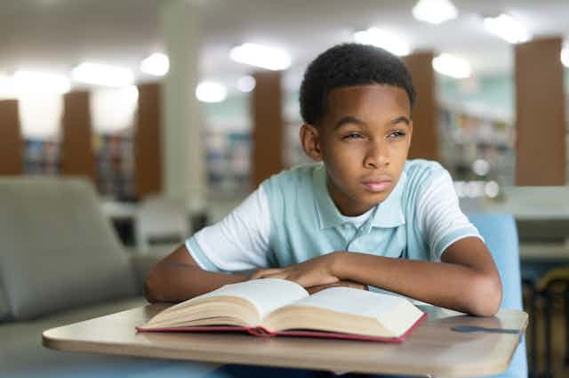 A boy sits at a desk in a library with an open book. He is looking off to his left.