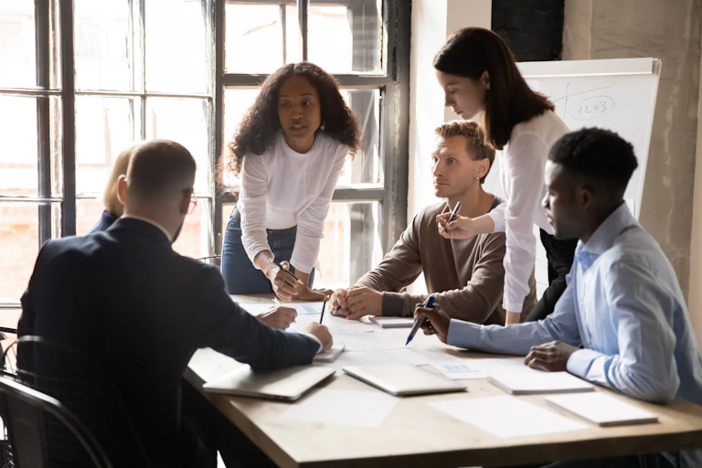 A group of visually diverse individuals having a conversation around a table in front of a whiteboard