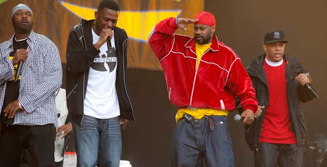 Four men seen performing on stage with microphones, one in a t-shirt that says 'Wu-Tang'