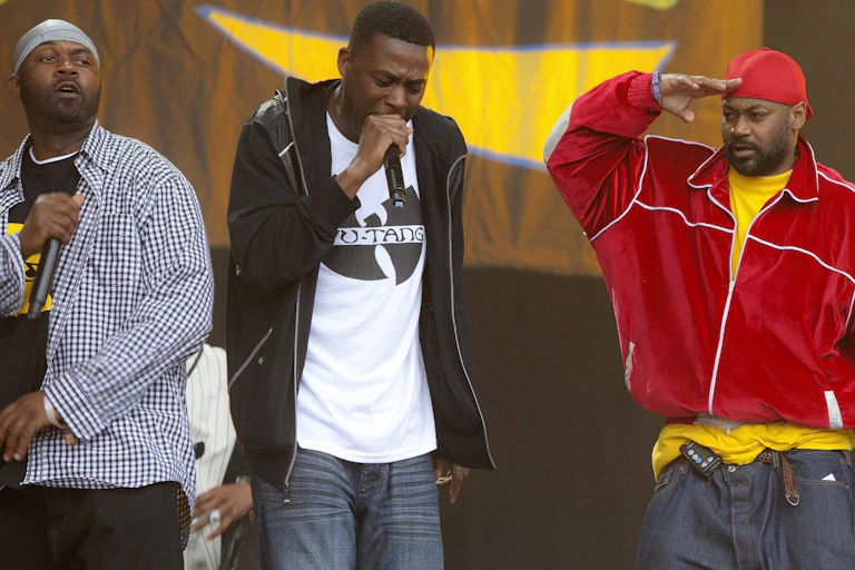 Four men seen performing on stage with microphones, one in a t-shirt that says 'Wu-Tang'