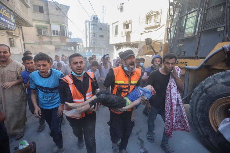 Two men wearing orange vests carry the body of a dead boy. They are surrounded by other people who are yelling and crying.