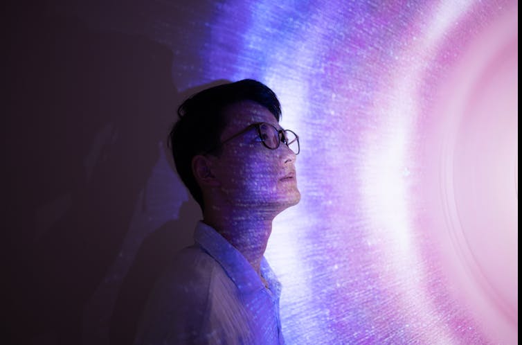 Un hombre asiático con gafas mira seriamente al espacio, de pie sobre un fondo holográfico en tonos de rosa y azul.