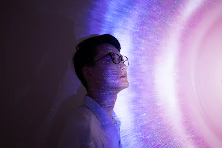 An Asian man wearing glasses stares seriously into space, standing against a holographic background in shades of pink and blue.