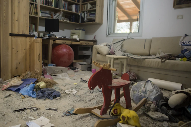 A living room with children's toys and a couch sits partially destroyed and covered in dust and rubble.
