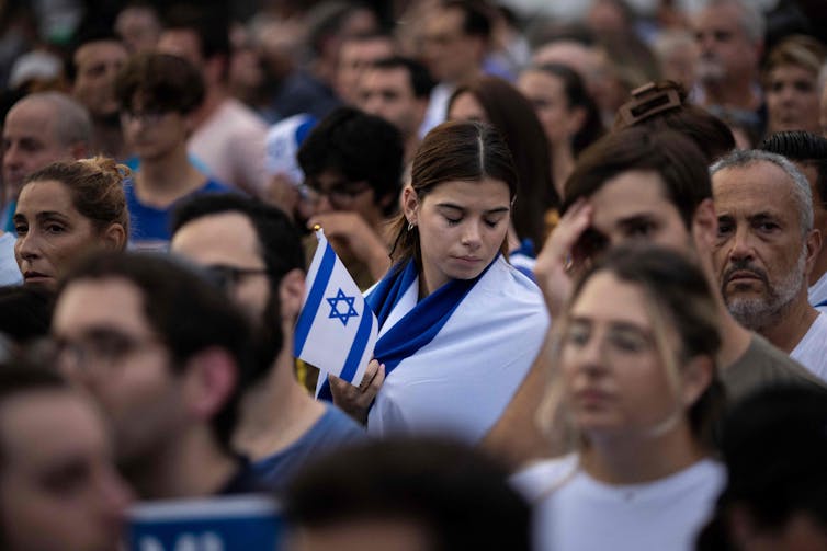 A crowd of people stand together, with one woman turning her face downward while holding an Israeli flag.