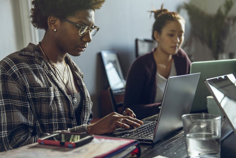 Two young women, one Black and one Asian, sit at a table together as they work on two laptops.