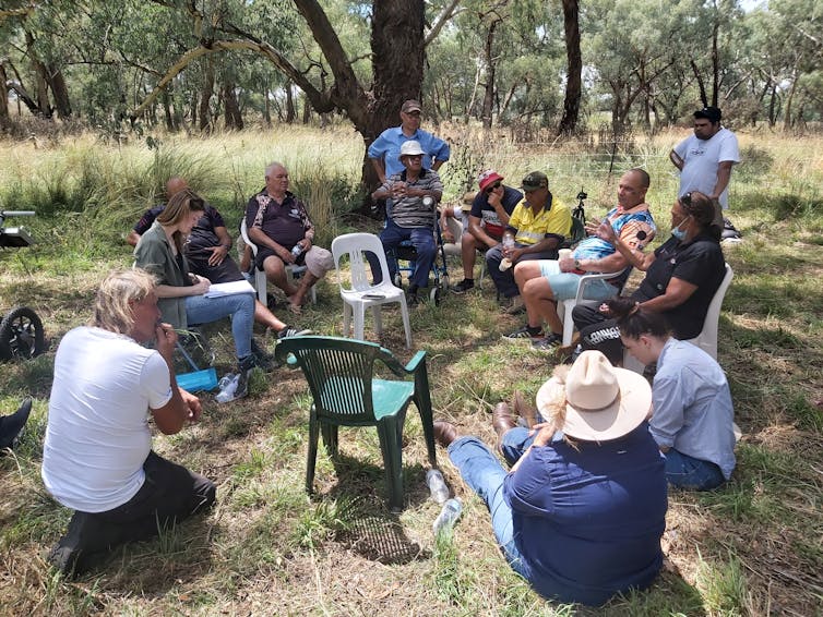 A group of people in discussion, sitting and standing in a circle outdoors.