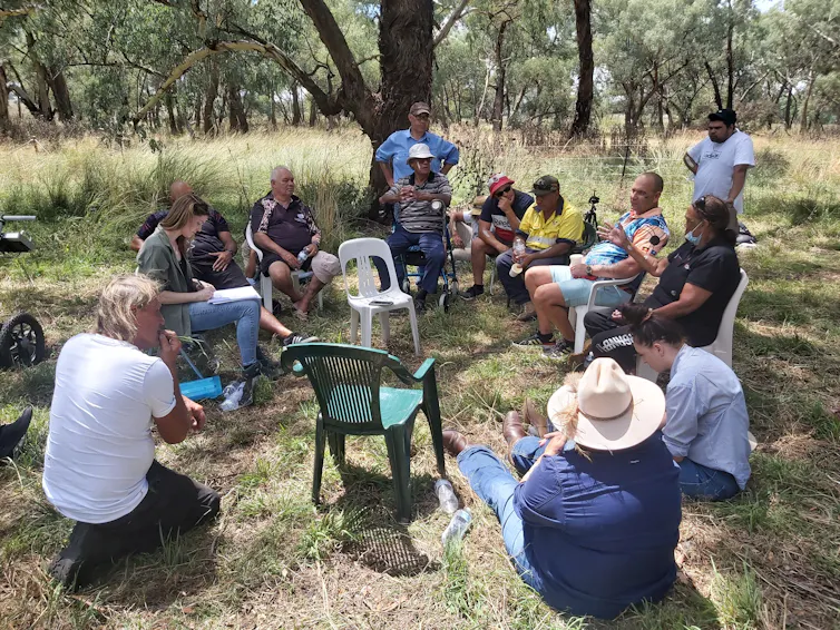 A group of people in discussion, sitting and standing in a circle outdoors.