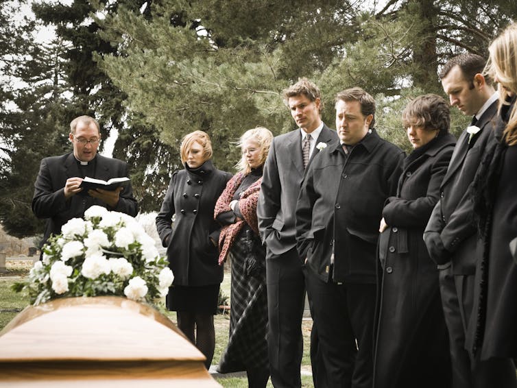 Seven people in a line, wearing black, stand near a coffin outside as another man with glasses reads from a book.