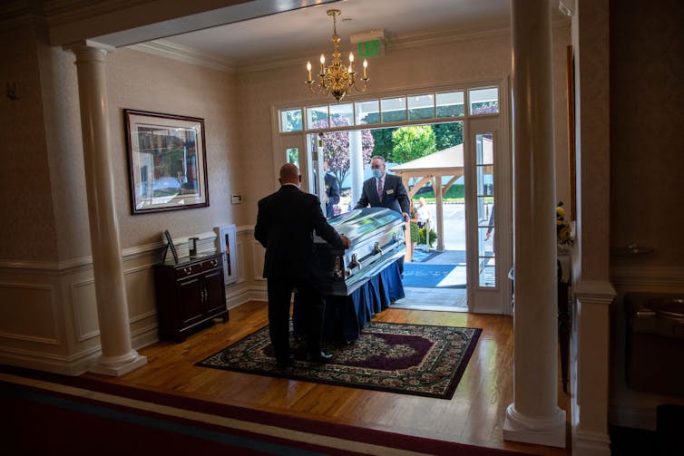 Two men in suits wheel a casket through a hallway and a door into the outdoors.