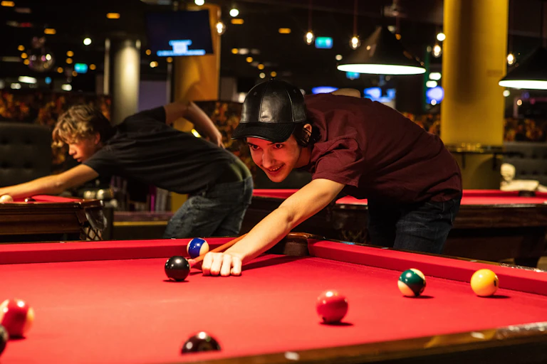 A young man lines up his cue stick to take a shot on a red velvet pool table in a darkened room.