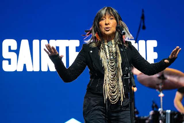 Buffy Sainte-Marie spreads her arms while singing on a stage.