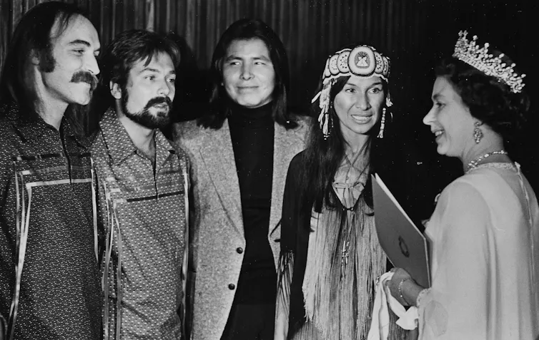 Three men are on Buffy Sainte-Marie's right as the Queen speaks to them.
