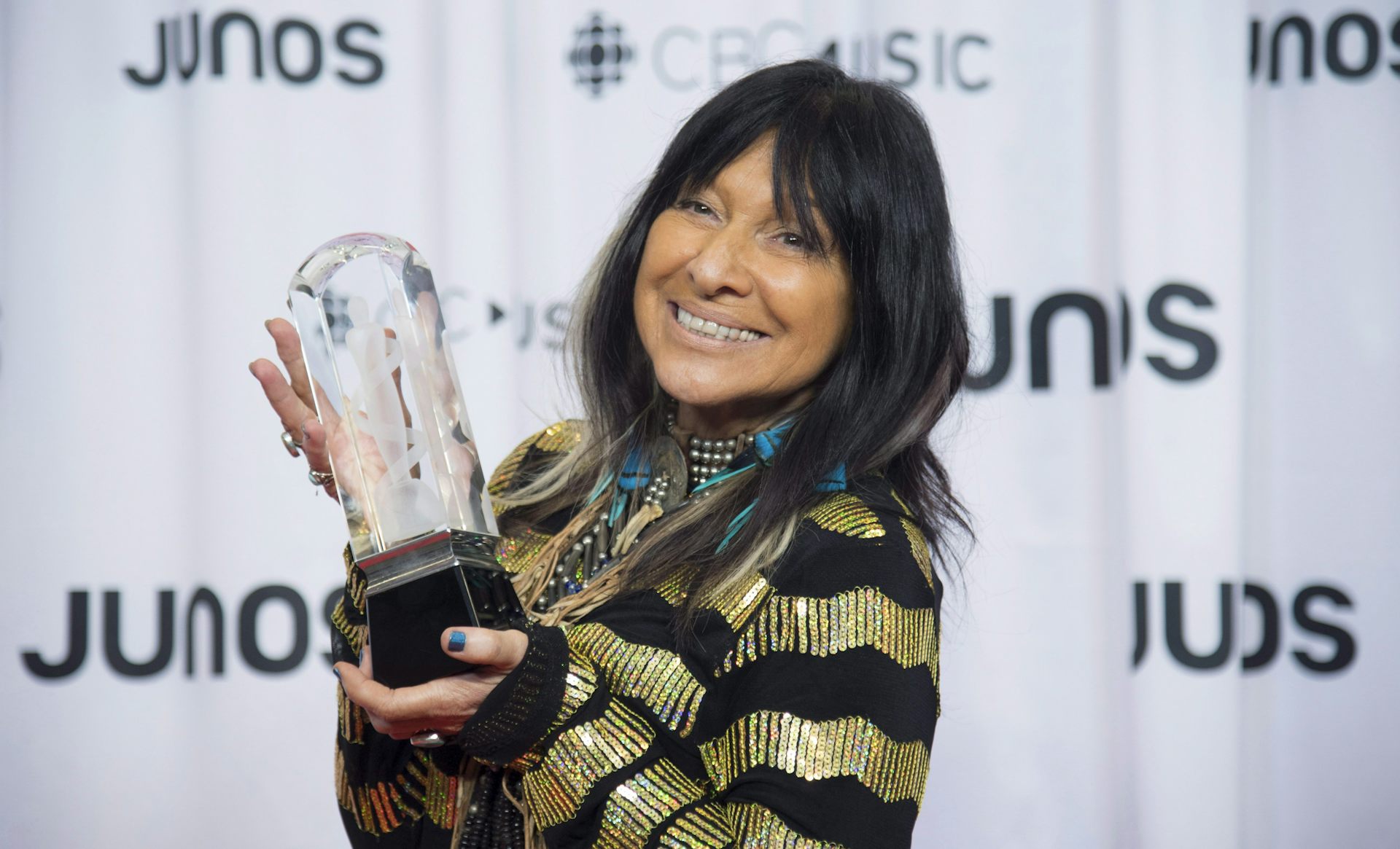 Buffy Sainte-Marie holds up an award in front of a curtain with the word Junos on it