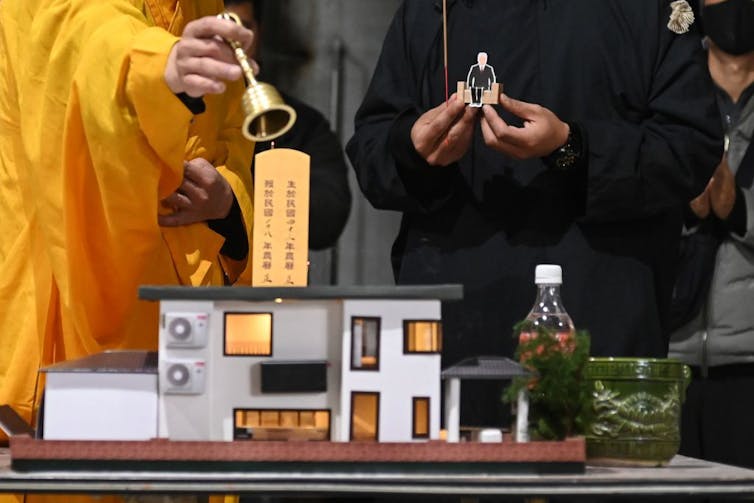 A man in a yellow monk's robe and someone wearing black stand behind what looks like a dollhouse, as the monk rings a bell.