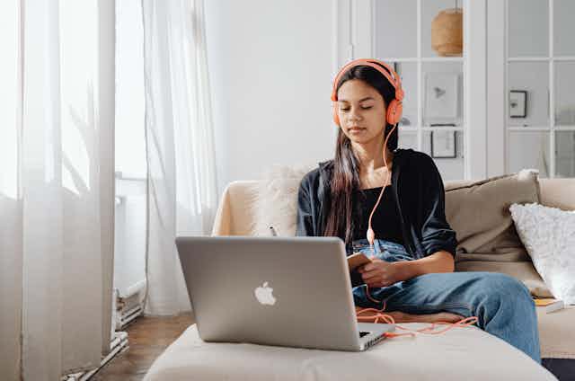 A young woman sits on a couch with a laptop and book, with headphones plugged in. 