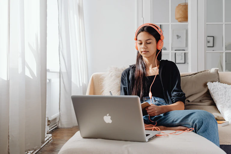 A young woman sits on a couch with a laptop and book, with headphones plugged in.