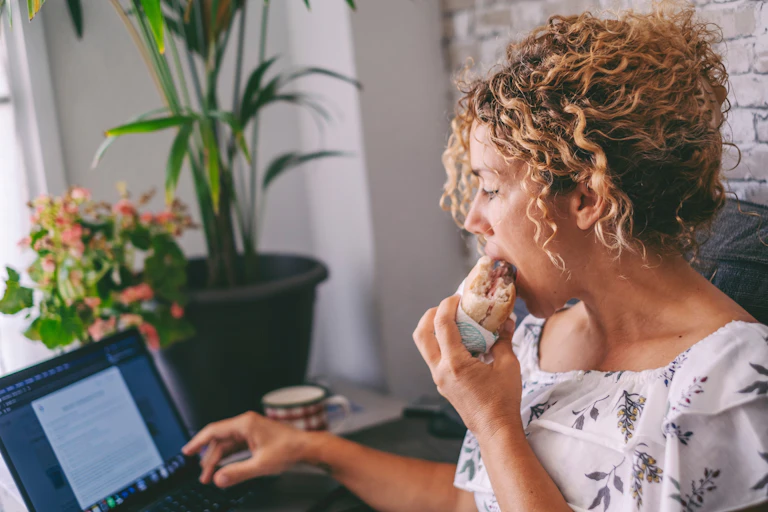 woman sits at laptop eating ham sandwich