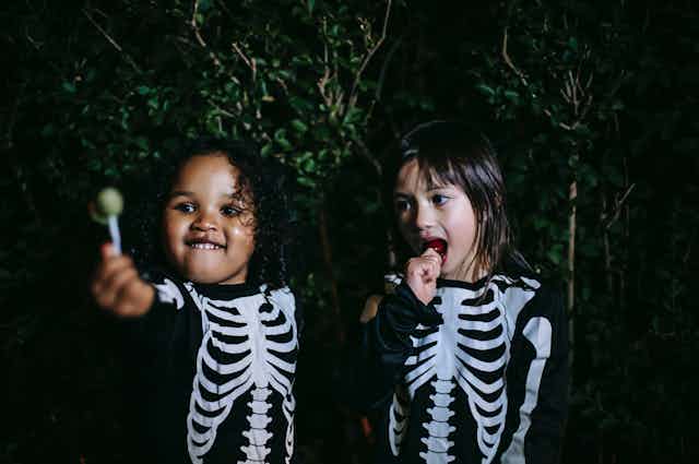 Two young children in skeleton costumes sit eating lollipops. 