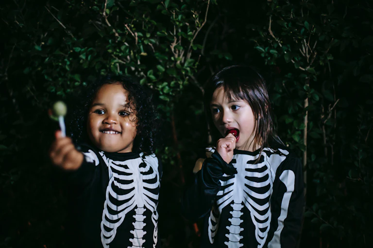 Two young children in skeleton costumes sit eating lollipops.