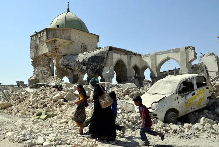 Mosul, Iraq, 2017: a family stands outside the ruins of a building with a wrecked car buried in the rubble.
