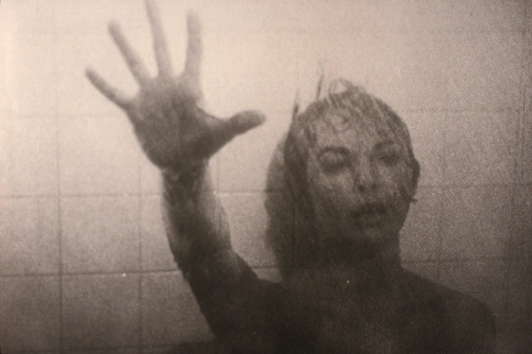 Escena de Psicosis en la que una mujer extiende el brazo buscando ayuda mientras su cuerpo se desliza por las baldosas de la pared.