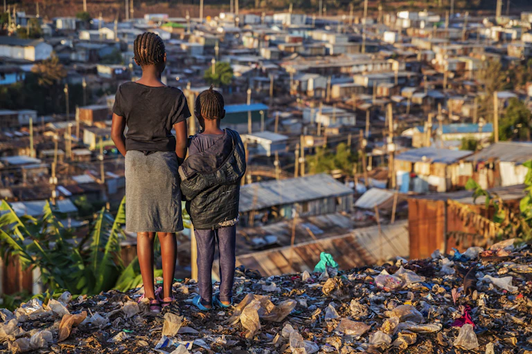 Two girls standing on a small cliffside littered with plastic bags and looking down at several makeshift homes made with iron sheets.